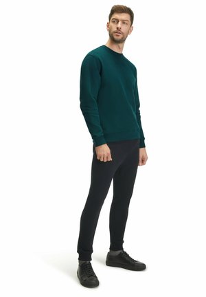 Donkergroen sweatshirt met ronde hals en geribbelde manchetten, gecombineerd met zwarte slim-fit broek en zwarte veterschoenen, tegen een witte achtergrond.