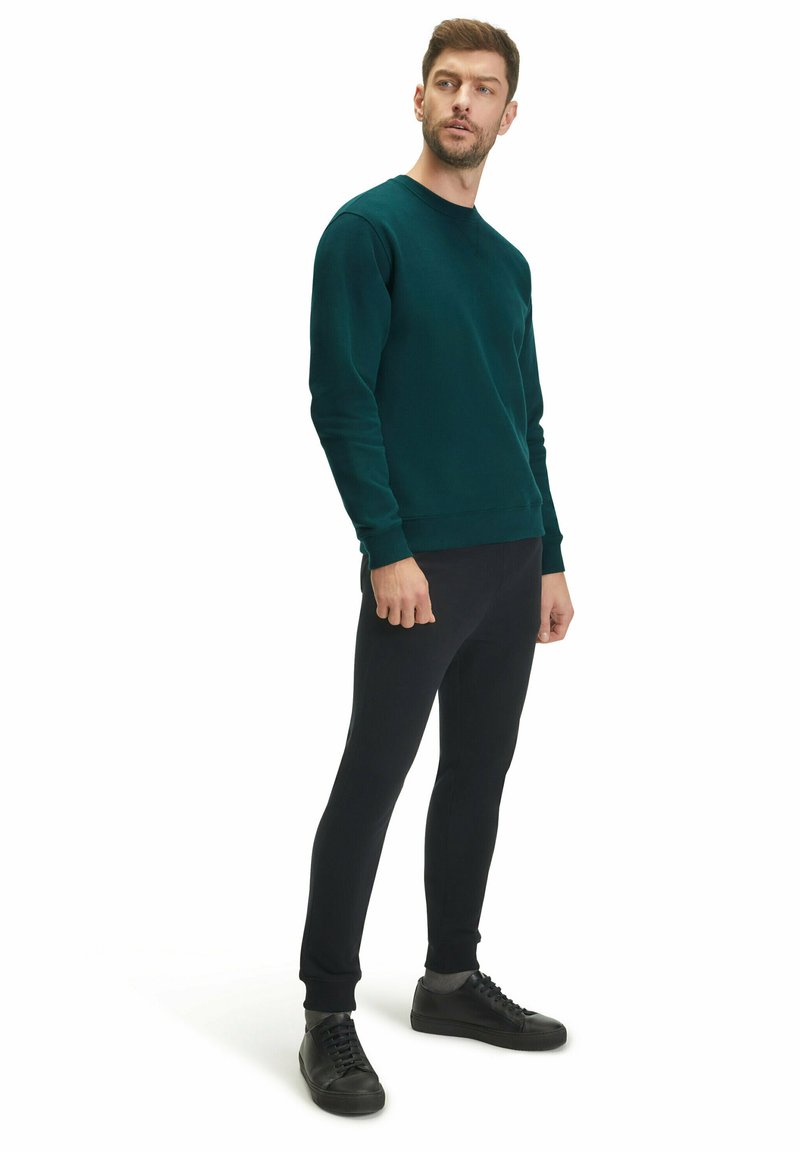 Felpa verde scuro a girocollo con maniche a costine, abbinata a pantaloni neri slim-fit e sneakers nere con lacci, su uno sfondo bianco.
