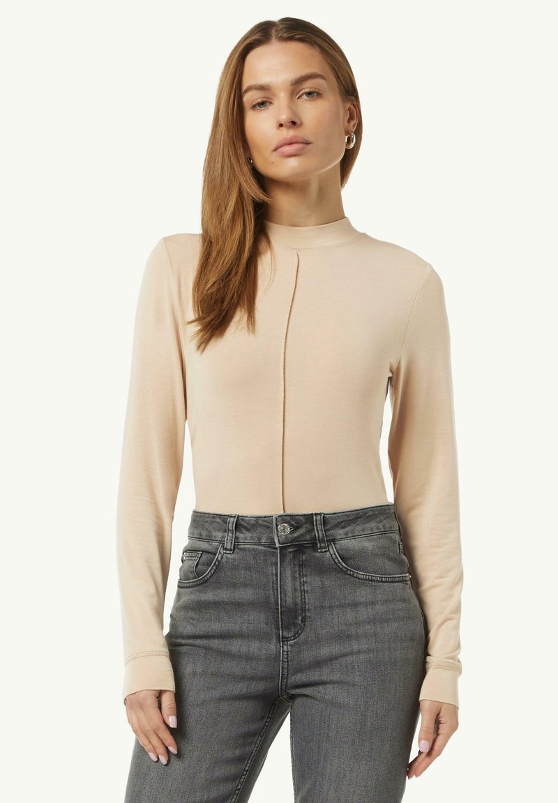 Beiger Langarm-Body mit hohem Kragen und vertikaler Nahtdetails, kombiniert mit hochgeschnittenen dunkelgrauen Jeans. Glatte Stofftextur.
