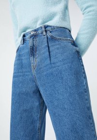 Jeans de mezclilla azul de tiro alto con un ajuste relajado, que presentan un desvanecimiento sutil, costuras de cobre y cierre de botón. Suéter texturizado azul claro encima.