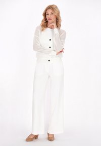 Weißer Netzcardigan mit schwarzen Knöpfen über einem weißen Oberteil, kombiniert mit weißen weit geschnittenen Hosen und beigefarbenen High-Heels. Glatte Textur überall.