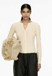 Helles Beige, gerippter Langarm-Cardigan mit V-Ausschnitt und Knopfschluss. Das Modell hält eine plüschige, mit Fell besetzte Tasche in einem helleren Farbton. Schwarze Hose.