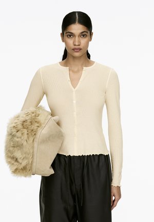 Helles Beige, gerippter Langarm-Cardigan mit V-Ausschnitt und Knopfschluss. Das Modell hält eine plüschige, mit Fell besetzte Tasche in einem helleren Farbton. Schwarze Hose.