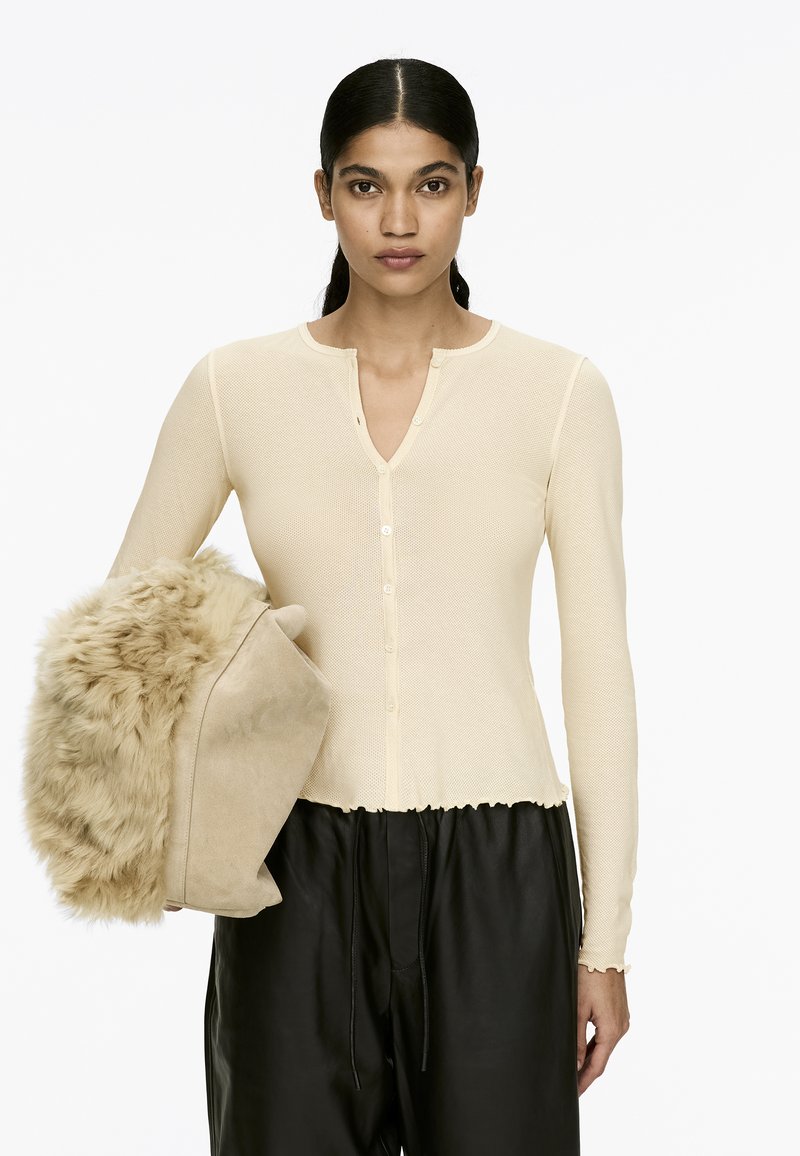 Helles Beige, gerippter Langarm-Cardigan mit V-Ausschnitt und Knopfschluss. Das Modell hält eine plüschige, mit Fell besetzte Tasche in einem helleren Farbton. Schwarze Hose.