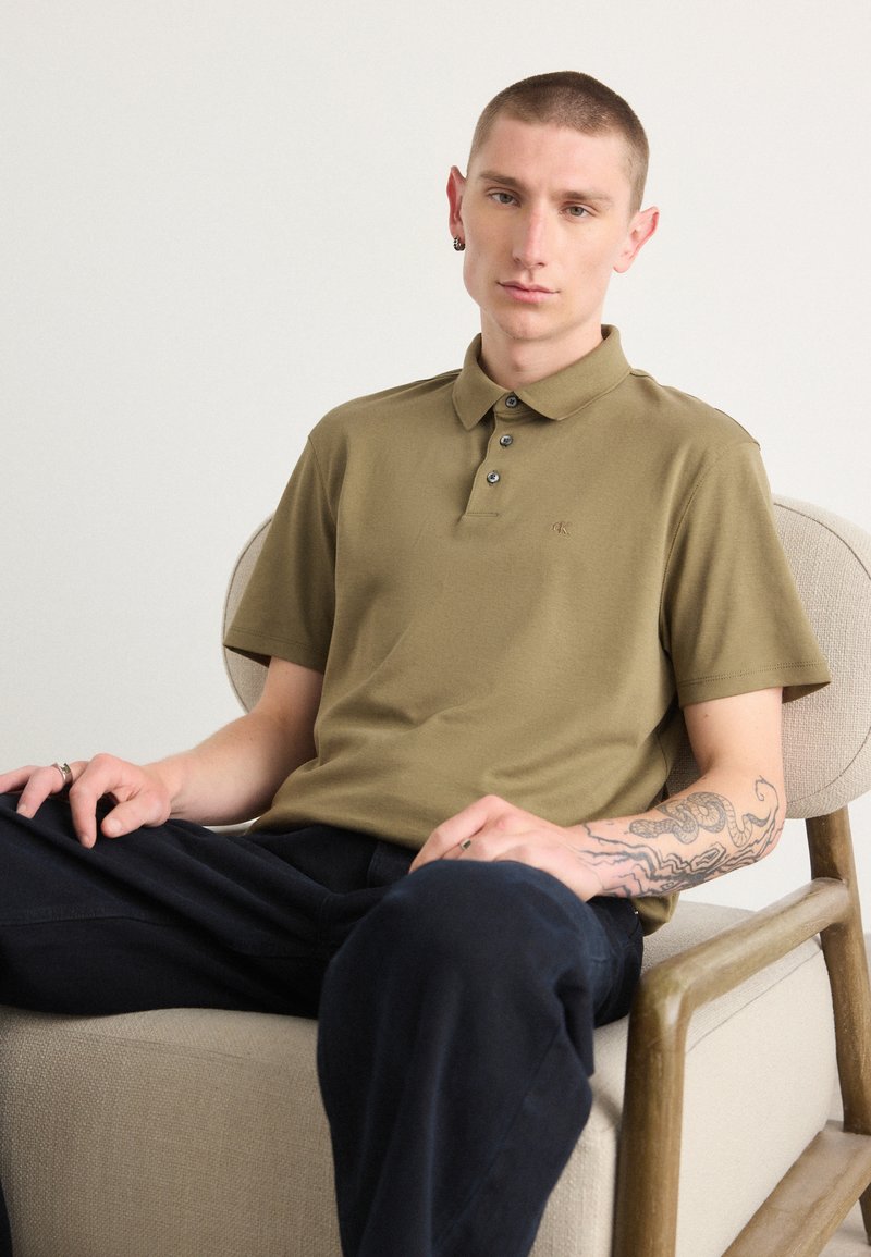 Polo shirt vert olive à manches courtes, avec un col à trois boutons et un tissu texturé. Porté avec un pantalon sombre, présentant un tatouage visible sur le bras.