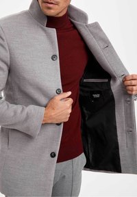 Grauer Wollmantel mit Stehkragen, fünf schwarzen Knöpfen und glattem schwarzem Innenfutter, getragen über einem burgunderroten Rollkragenpullover.