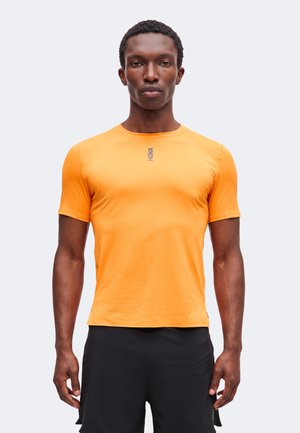 Uomo con capelli corti e ricci che indossa una maglietta sportiva arancione a maniche corte e pantaloncini neri, in piedi davanti a uno sfondo bianco semplice.