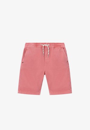Enfant souriant aux cheveux bouclés portant un t-shirt rayé bordeaux et gris, un short corail, des chaussettes dépareillées et des baskets beiges sur fond beige.