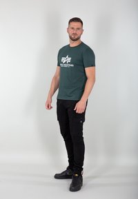 Tmavě zelené tričko s bílým logem, zkombinované s úzkými černými cargo kalhotami s kapsami. Outfit doplňují černé tenisky.