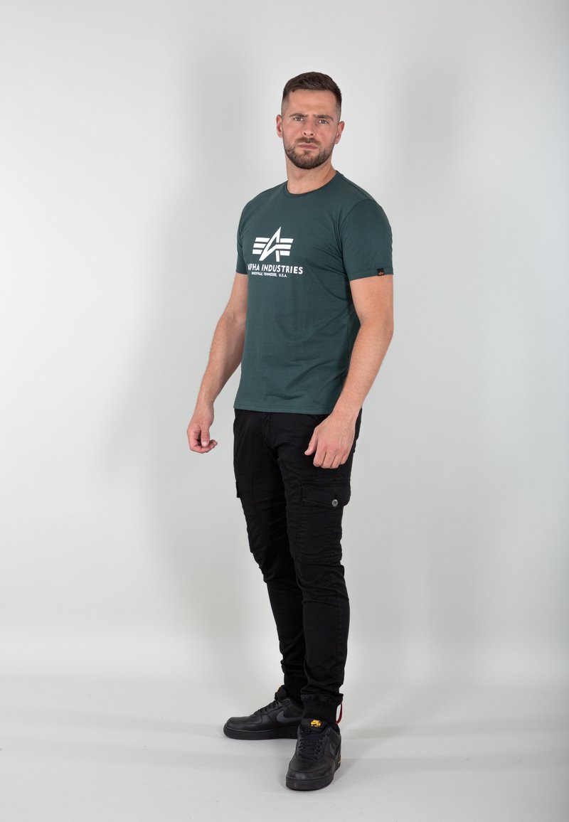 Dunkelgrünes T-Shirt mit einem weißen Logo, kombiniert mit schmal geschnittenen schwarzen Cargo-Hosen mit Taschen. Schwarze Sneakers vervollständigen das Outfit.