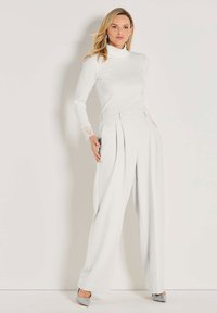 Vrouw in een witte coltrui met kanten manchetten, hoge taille wijde broek en grijze puntige hakken, staande tegen een witte muur.