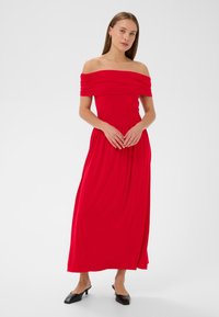 Robe maxi rouge décolletée avec un col froncé, texture lisse, bustier ajusté et jupe fluide. Portée avec des mules à talons noirs.