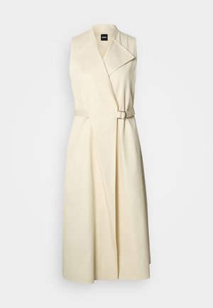 Μακρύ μπεζ παλτό τύπου καπαρντίνα χωρίς μανίκια, με φαρδιές πέτρες και ταιριαστή ζώνη με αγκράφα στη μέση.