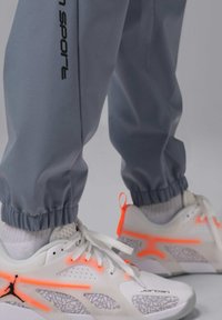 Białe buty sportowe z neonowo-pomarańczowymi akcentami oraz szare, teksturowane spodnie dresowe z elastycznymi ściągaczami i czarnym logo po boku.