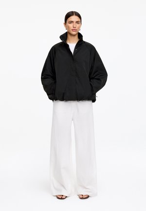 Vrouw draagt een zwart oversized jack met hoge kraag, witte wijde broekspijpen en zwarte sandalen met open teen, staande tegen een witte achtergrond.