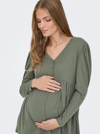 Zwangere vrouw in een lang mouw, olijfgroene jurk die voorzichtig haar buik vastneemt en met een zachte, tevreden uitdrukking naar beneden kijkt.