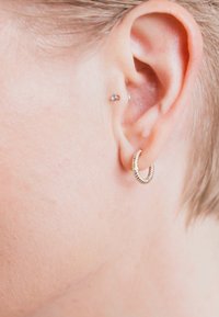 Zlatý kruhový náušník s textúrovaným vzorom, umiestnený v prepichnutom uchu, doplnený malým strieborným náušníkom nad ním.
