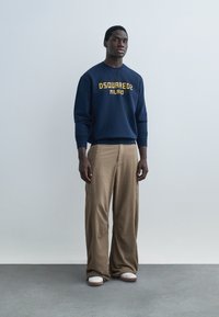 Man draagt marineblauwe trui met tekst "DSQUARED2 MILANO", losse beige broek en witte sneakers, staand tegen een effen achtergrond.