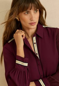 Bordo värvi pluus nööpidega esiküljel, kontrastsete triibuliste detailidega krae ja mansettide juures ning plisseeritud varrukatega. Sile kangapind.