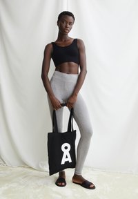 Zwart cropped top, grijze leggings en zwarte draagtas met wit logo. Model staat tegen een lichte achtergrond en draagt zwarte slides.