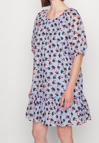 Jasnoniebieska luźna sukienka z czarno-czerwonym kwiatowym wzorem, bufiastymi rękawami i falbaną na dole, noszona przez stojącą osobę.