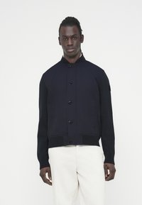 Στρατιωτικό μπουφάν ναυτικού με πλεκτά μανίκια, όρθιο γιακά και κούμπωμα με κουμπιά. Διαθέτει σώμα από λείο ύφασμα και ριμπ τελείωμα και μανσέτες.