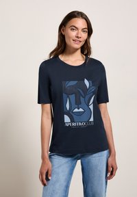 T-shirt bleu marine à manches courtes, ornée d'un design graphique d'un visage et de feuilles dans des tons de bleu plus clairs. Le texte en bas indique "APERTIVO CLUB CÔTE AMALFITE".