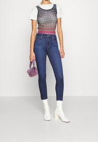 Top de punto sin mangas en un patrón multicolor, superpuesto a una camiseta blanca, combinado con pantalones ajustados de mezclilla azul oscuro y botas blancas hasta el tobillo.