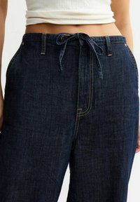 Pantalons en denim bleu foncé avec une taille à cordon, présentant des poches avant standards et des coutures contrastantes le long des seams.