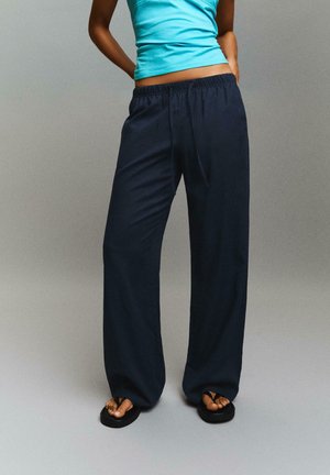 Nő laza sötét tengerészkék nadrágot, fekete platform szandált és élénk kék ujjatlan felsőt visel, zsebre tett kézzel áll.