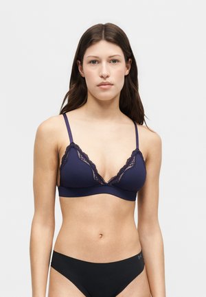 Femme portant un soutien-gorge bleu marine bordé de dentelle et une culotte noire, debout devant un fond gris clair uni, face à la caméra.