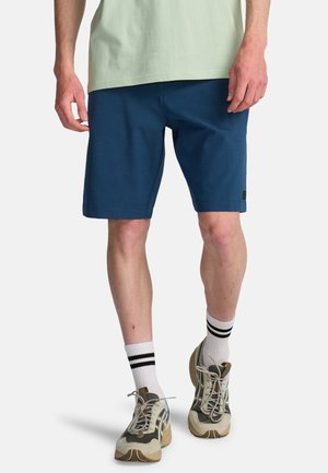 Man die marineblauwe korte broek, witte sokken met zwarte strepen en beige sportschoenen draagt, staand tegen een witte achtergrond.