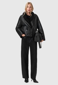 Zwarte leren dubbelbreasted jas met een brede sjawlcollar, gedragen over losse zwarte broeken en hoge hakken, met een kleine handtas.