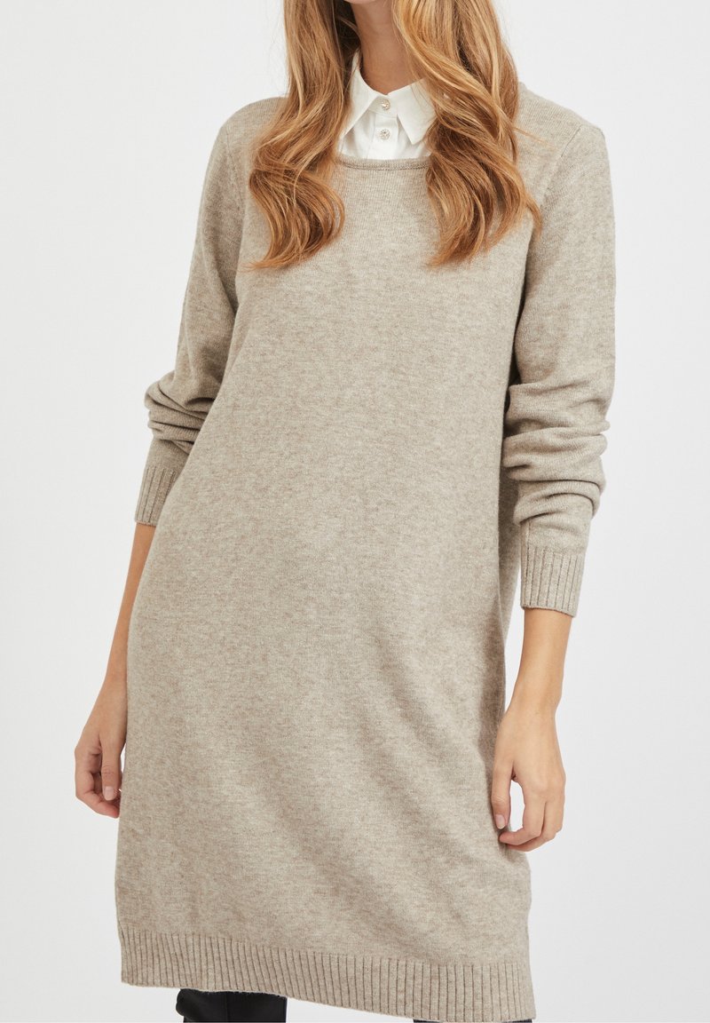 Robe-pull beige en maille avec poignets et ourlet côtelés. Encolure ronde, manches longues et coupe décontractée. Le matériau semble doux avec une texture subtile.