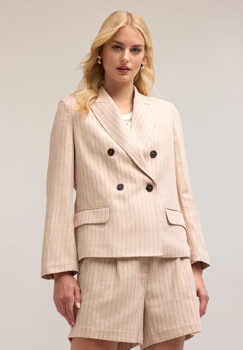 Blazer doppiopetto rosa chiaro con righe verticali, bottoni neri e due tasche frontali. Abbinato a pantaloni shorts a vita alta coordinati.