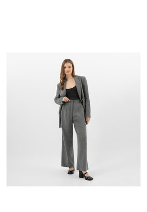 Kobieta w szarym garniturze z szerokimi spodniami i czarnym topem, stojąca na tle gładkiego, jasnego tła, ubrana w czarne buty na obcasie.