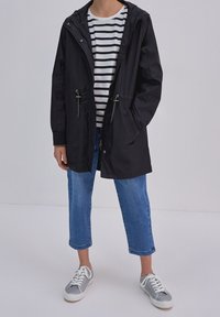 Zwarte waterdichte parka met capuchon, voorzien van voorkeursluitingen, twee zakken en verstelbare koorden, gecombineerd met een gestreept shirt en blauwe jeans.