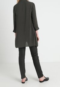 Camicia lunga bianca e nera a pois con maniche arrotolabili, abbinata a pantaloni neri e scarpe slip-on basse, su uno sfondo minimale.