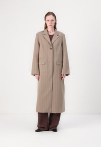 Brown striped long coat with a single button closure, two front pockets, notched lapels, and wide sleeves, worn over a dark brown outfit.