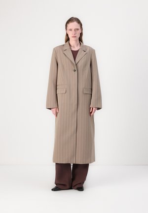 Cappotto lungo a righe marroni con chiusura a un solo bottone, due tasche frontali, rever a punta e maniche larghe, indossato sopra un abito marrone scuro.