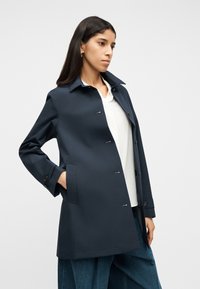 Γυναίκα που φοράει σκούρο μπλε παλτό πάνω από λευκό πουκάμισο και φαρδύ μπλε παντελόνι, στέκεται με το ένα χέρι στην τσέπη μπροστά σε απλό φόντο.