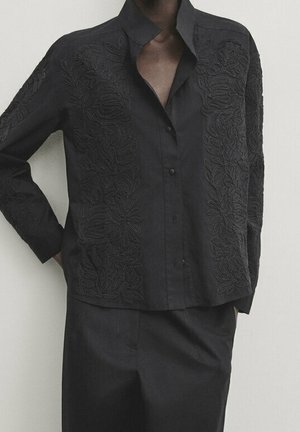 Zwarte blouse met lange mouwen en knopen, voorzien van bloemrijke borduurwerk, gedragen met een bijpassende zwarte rok, handen van het model in de zakken, staand tegen een effen achtergrond.