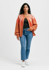 Oranssin värinen neulecardigan, jossa on valkoisia geometrisiä kuvioita. Siinä on pyöreä kaula-aukko, yli-iso malli ja väljät hihat. Cardigan on puettu mustan paidan ja sinisten farkkujen päälle.
