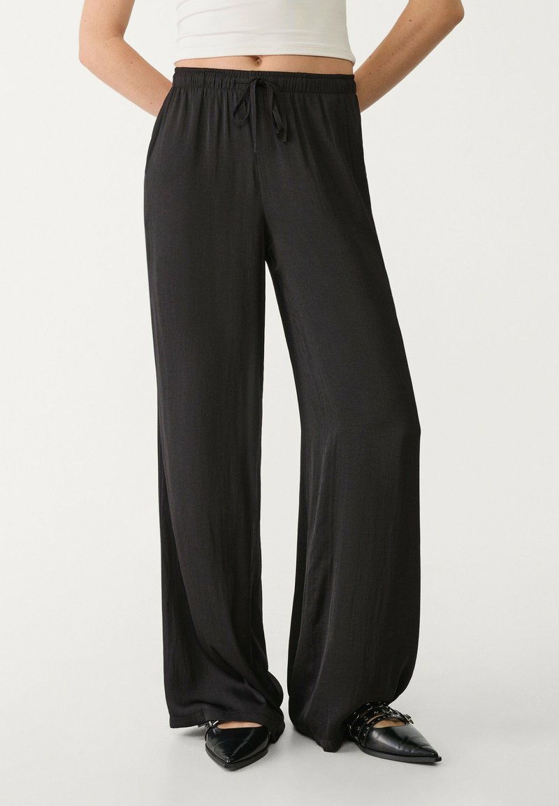 Schwarze, locker sitzende Hosen mit einem elastischen Bund und Kordelzug. Hergestellt aus einem glatten, leichten Stoff.