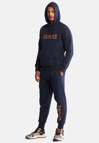 Ensemble de sweat à capuche et pantalon de jogging bleu marine en coton, avec une poche avant, un logo Timberland orange et des poignets côtelés. Les chaussures sont beiges et noires.