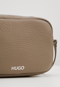 Hnědá kožená taška s texturovaným povrchem, se bílým logem "HUGO". Má obdélníkový tvar a štíhlý, strukturovaný design.