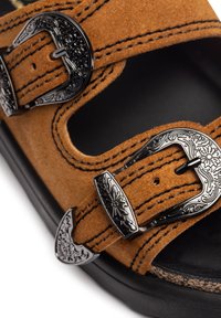 Sandale en daim marron avec deux boucles décoratives en métal noir et des coutures contrastées. Elle possède une ouverture au niveau des orteils et une semelle intérieure ergonomique pour le confort.