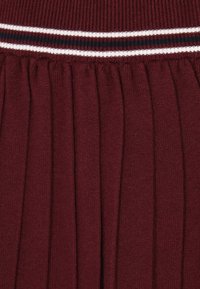 Burgundy red, redőzött szoknya texturált kötött anyagból. Jellemzője a bordázott derekú rész, amelyet fehér és sötétkék csíkok díszítenek.