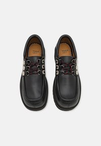 Zapatos de cuero negros con una forma redondeada, que presentan herrajes plateados y cordones rojos y negros. La suela tiene un diseño texturizado.