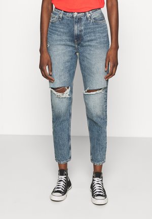 Personne portant un jean déchiré taille haute bleu clair associé à des baskets montantes noires et un haut rouge, debout sur un sol blanc uni.